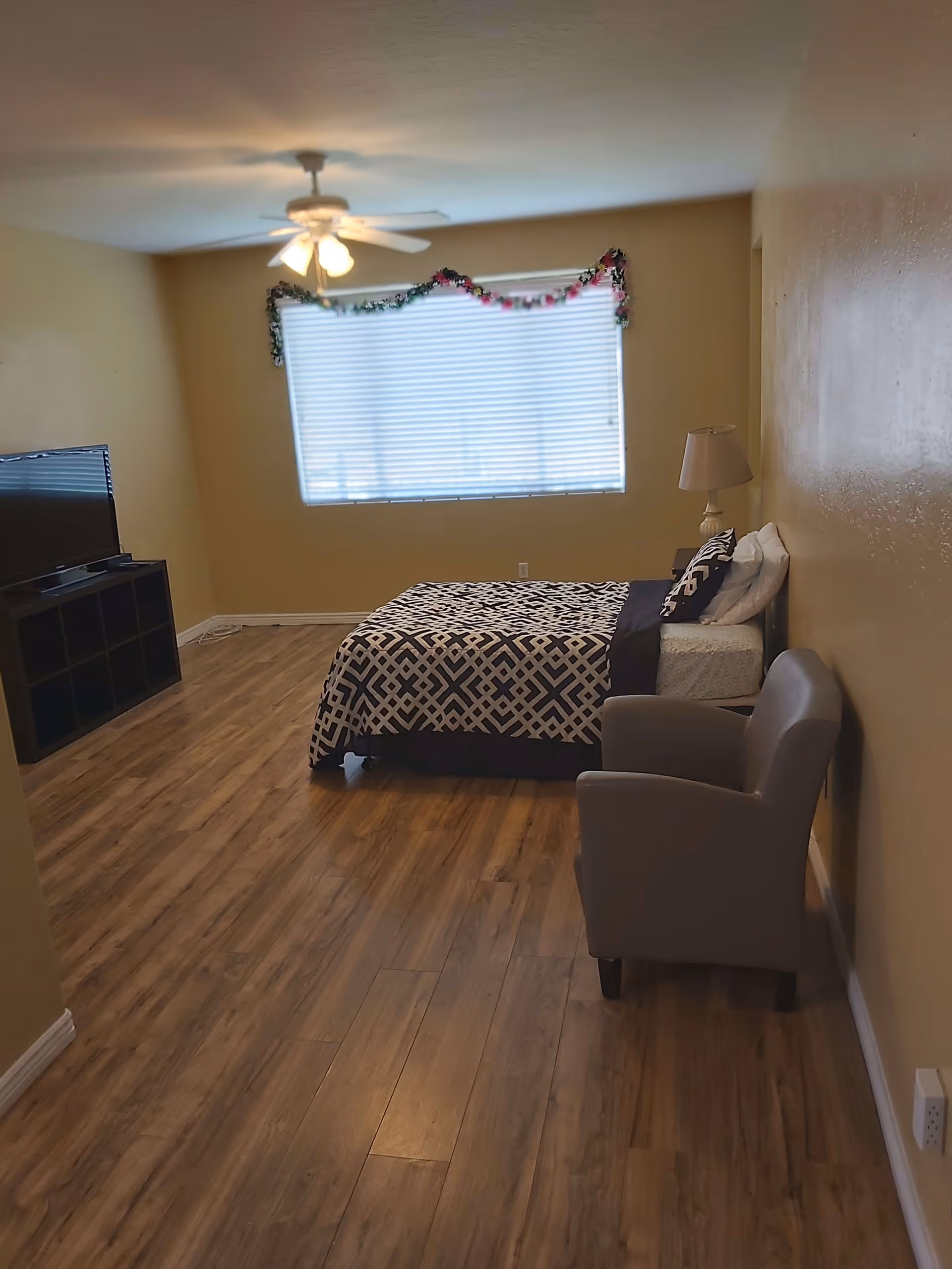 A bedroom with wooden flooring, a bed with a patterned black and white bedspread, a gray armchair, a lamp on a nightstand, a ceiling fan with lights, a large window with blinds and garland decoration, and a TV on a black stand.