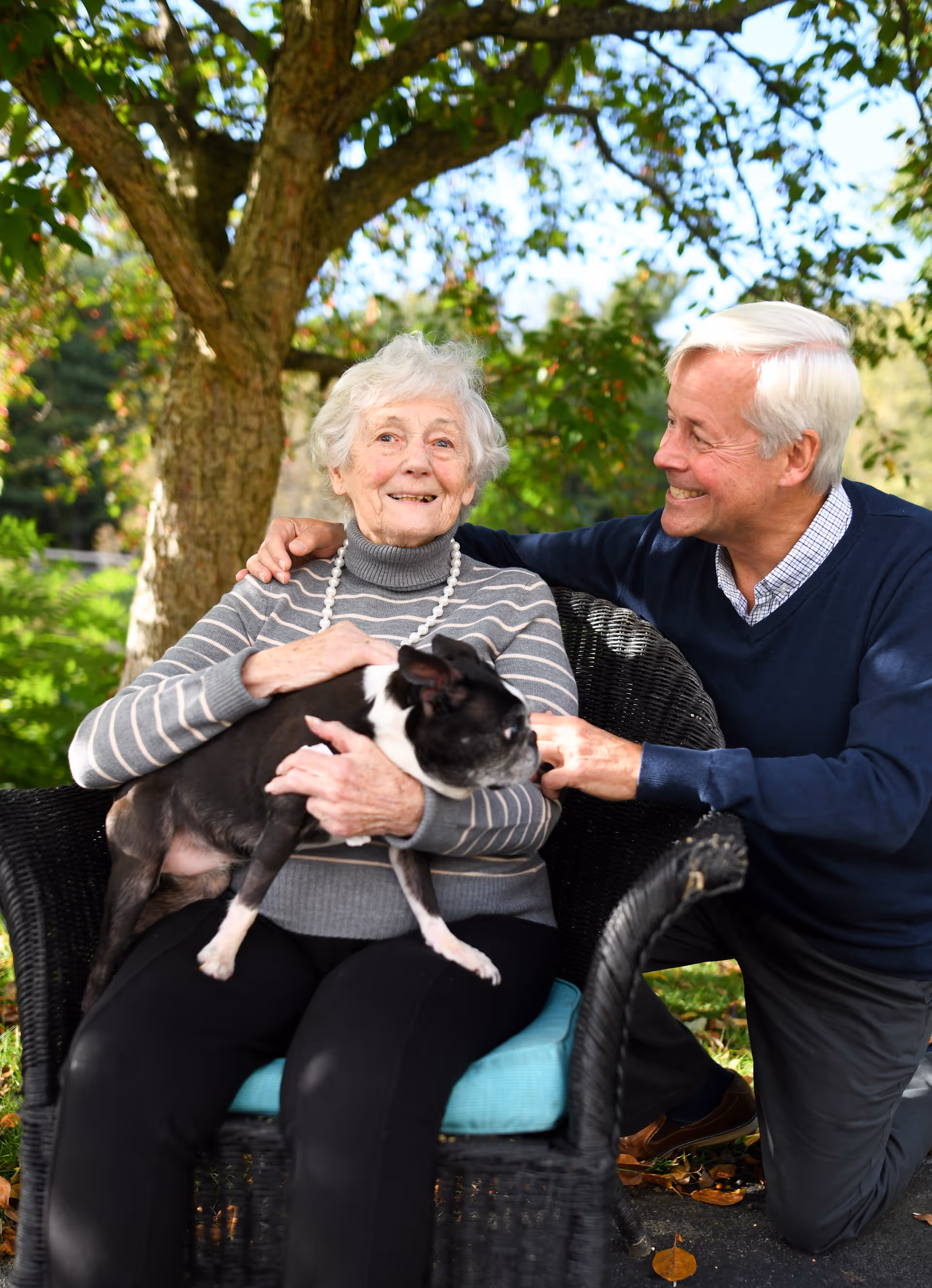 An elderly woman sitting on a wicker chair outdoors holding a small black and white dog, with an elderly man kneeling beside her smiling and resting his arm on her shoulder. They are surrounded by greenery and a tree in the background.