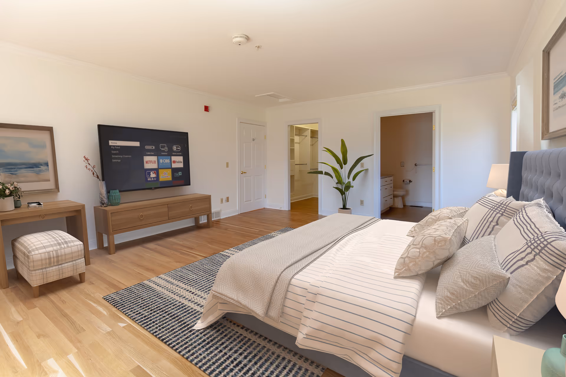 A spacious bedroom with a large bed adorned with multiple pillows and a striped bedspread. The room features hardwood floors, a patterned area rug, a flat-screen TV mounted on the wall above a wooden console, a small desk with a plaid cushioned stool, and a potted plant near two open doorways leading to a walk-in closet and a bathroom. The walls are painted white, and there is framed artwork on the walls.