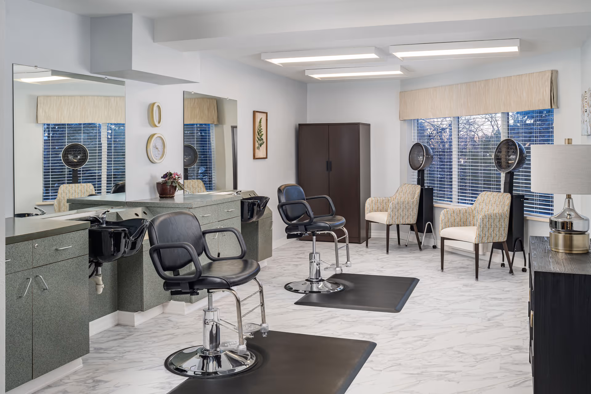 Interior view of a senior living facility's hair salon with two black salon chairs in front of large mirrors and sinks. The room has a light color scheme with marble-patterned flooring, two beige patterned armchairs near the window, two hair dryers, a dark wooden cabinet, and a table lamp on a side table.