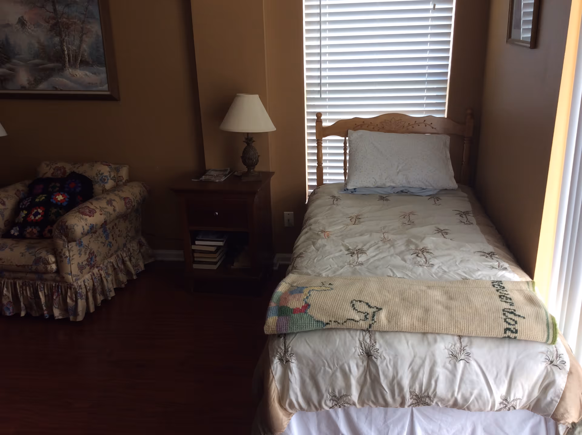 A small bedroom with a single wooden bed covered with a white bedspread featuring palm tree patterns. A folded blanket is placed at the foot of the bed. Next to the bed is a wooden nightstand with a decorative lamp and some books on the lower shelf. To the left of the nightstand is a floral-patterned armchair with a colorful crocheted pillow. The room has brown walls and a window with white blinds behind the bed.