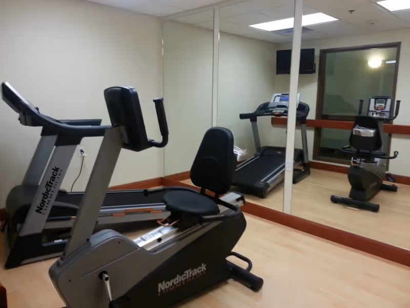 Small fitness room with exercise equipment including a recumbent bike, treadmill, and an upright bike. The room has light-colored walls, a large mirror covering one wall, and a window with a wooden ledge.