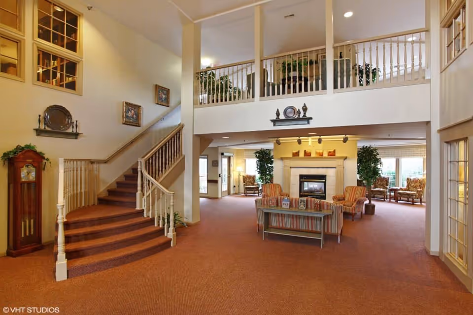 Spacious assisted living facility common area with a staircase on the left, a fireplace in the center surrounded by chairs and a sofa, and large windows letting in natural light. The room features warm carpeting and decorative plants, with a second-floor balcony overlooking the area.