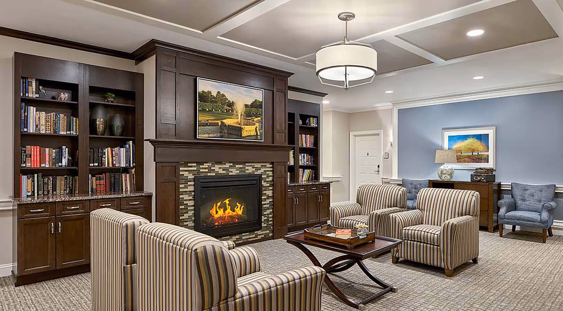 A cozy living room area with a lit fireplace surrounded by dark wood cabinetry and bookshelves. The room features four striped armchairs arranged around a wooden coffee table. The walls are painted in neutral tones with one accent wall in blue, decorated with framed artwork and a table lamp on a sideboard. The ceiling has recessed lighting and a modern chandelier.