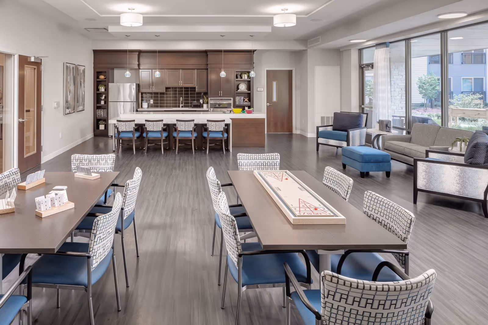 A spacious and modern common area in a senior living facility featuring a kitchen with dark wood cabinets, a refrigerator, and a long counter with five chairs. In the foreground, there are two tables with patterned chairs, one table has a shuffleboard game on it. To the right, there is a seating area with a sofa, armchairs, and an ottoman near large windows that let in natural light and show an outdoor garden area.