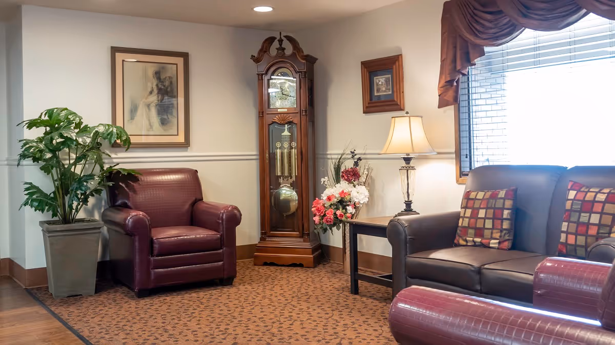 Comfortable sitting area with leather armchairs and a sofa, a grandfather clock, lamp, and plants beside a window.