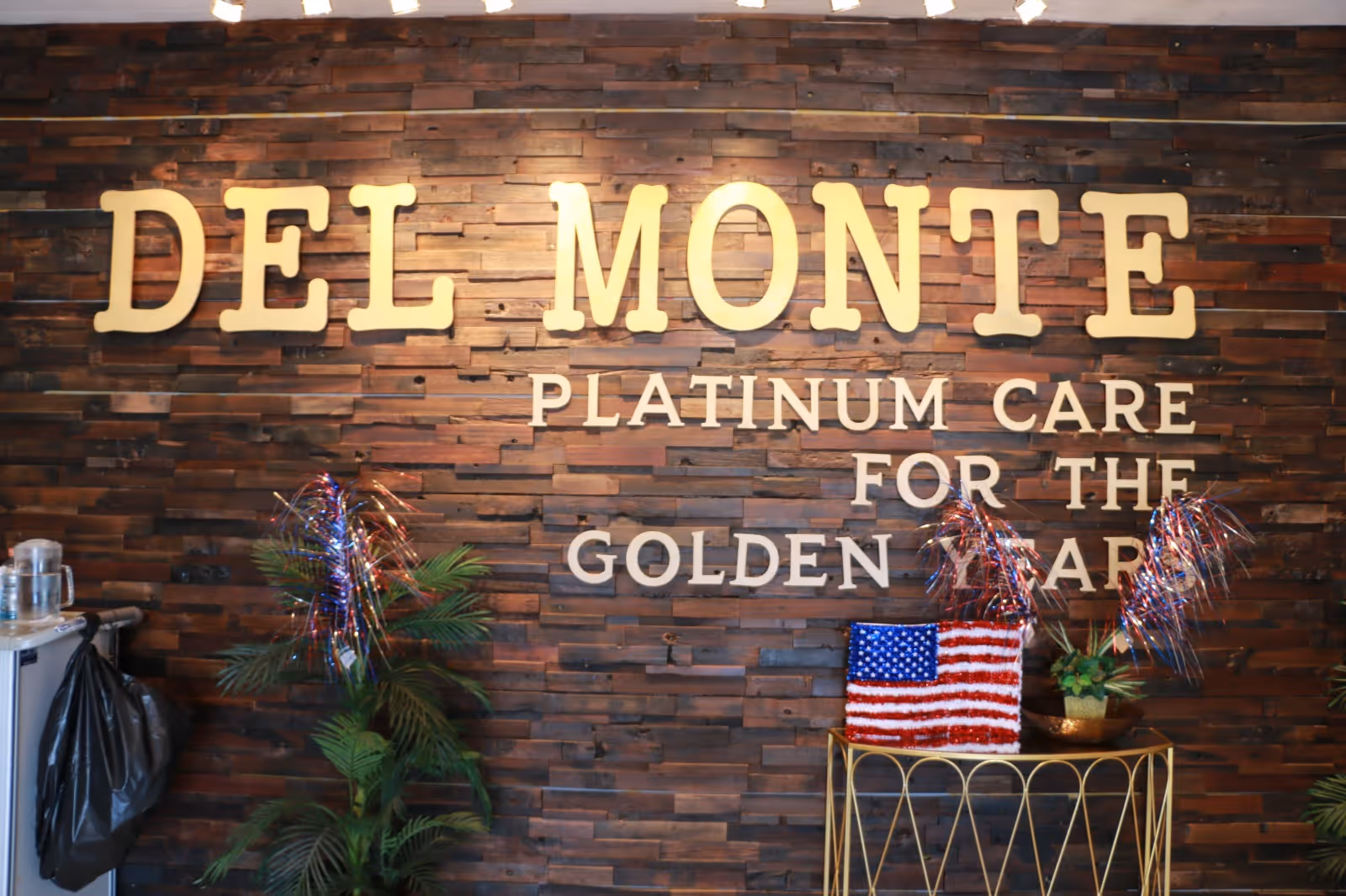 Wood-paneled interior wall sign reading 'DEL MONTE PLATINUM CARE FOR THE GOLDEN YEAR' with decorative plants and an American flag pillow on a small table.