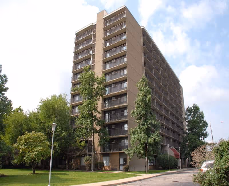 Exterior view of a multi-story senior living facility building named Carnegie Tower at Fairfax, surrounded by trees and greenery under a partly cloudy sky.