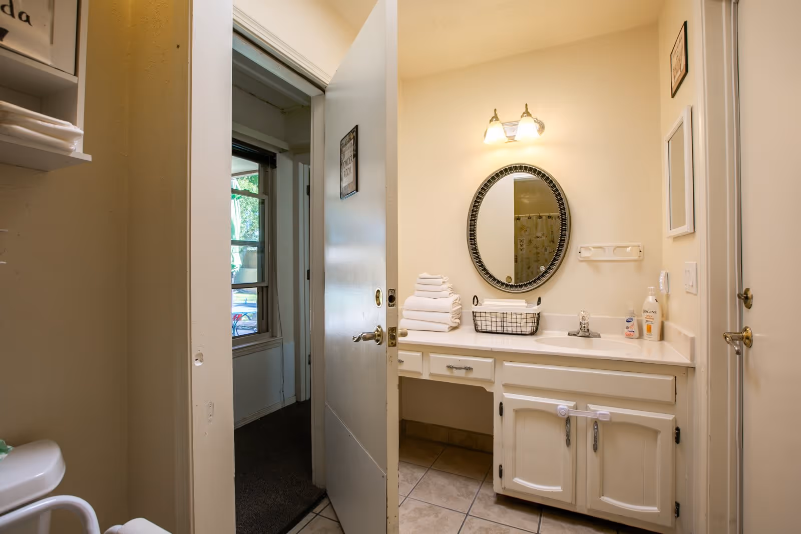 Interior view of a bathroom with a white vanity cabinet, a round mirror above the sink, a light fixture with two bulbs, and a stack of white towels next to a wire basket on the countertop. The bathroom door is open, showing a glimpse of an adjacent room with a window.