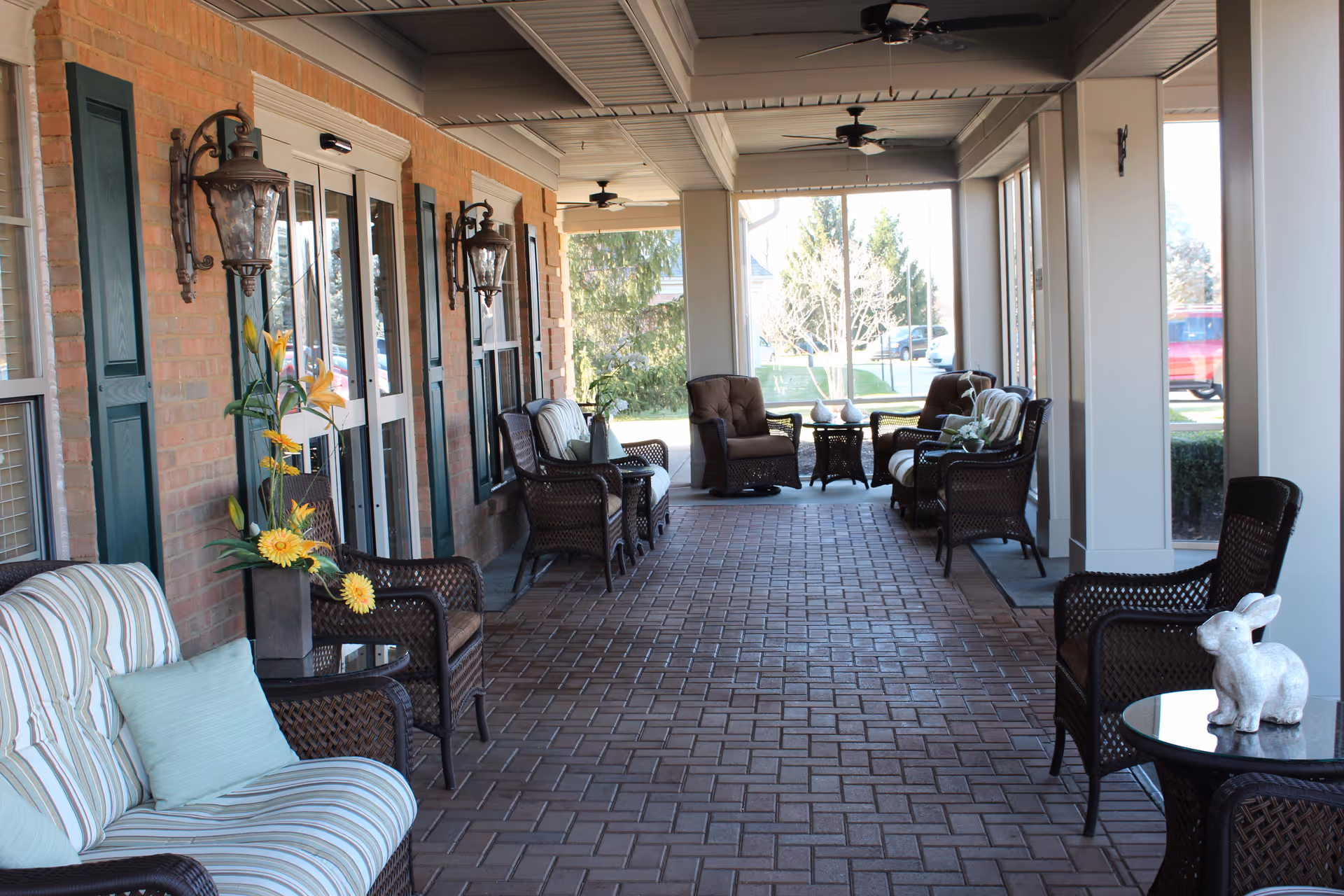 Covered outdoor patio area with brick flooring and multiple wicker chairs and tables arranged along the sides. The left side features a brick wall with windows and decorative wall-mounted lanterns. There are ceiling fans overhead and some decorative items like flowers and a small rabbit statue on the tables. The patio overlooks a green outdoor space with trees and parked cars visible in the background.