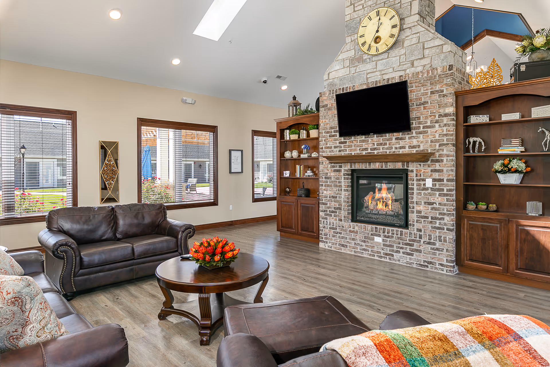 A cozy living room with leather sofas and a round wooden coffee table with a flower arrangement. The room features a large brick fireplace with a mounted TV above it, flanked by wooden built-in shelves with decorative items. Large windows allow natural light to brighten the space, and a colorful blanket is draped over one sofa.