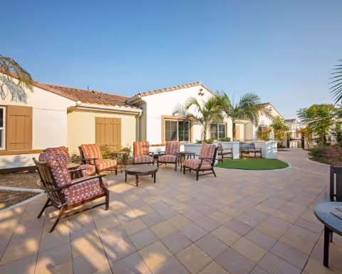 Sunlit outdoor courtyard featuring cushioned patio chairs and tables arranged on a tiled patio in front of a Mediterranean-style building with palm trees.