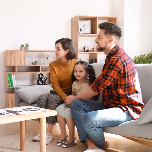 A family of three sitting on a light gray sofa in a modern living room. The man in a red plaid shirt and jeans is holding the hands of a young girl in a light dress, while a woman in a mustard sweater and checkered pants sits next to them. Behind them is a wooden shelving unit with decorative items and books.