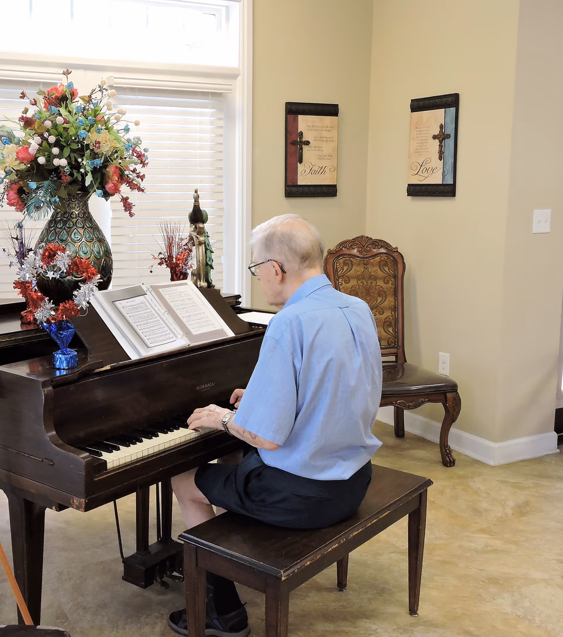 An elderly person seated at an upright piano playing from sheet music in a decorated living room area.