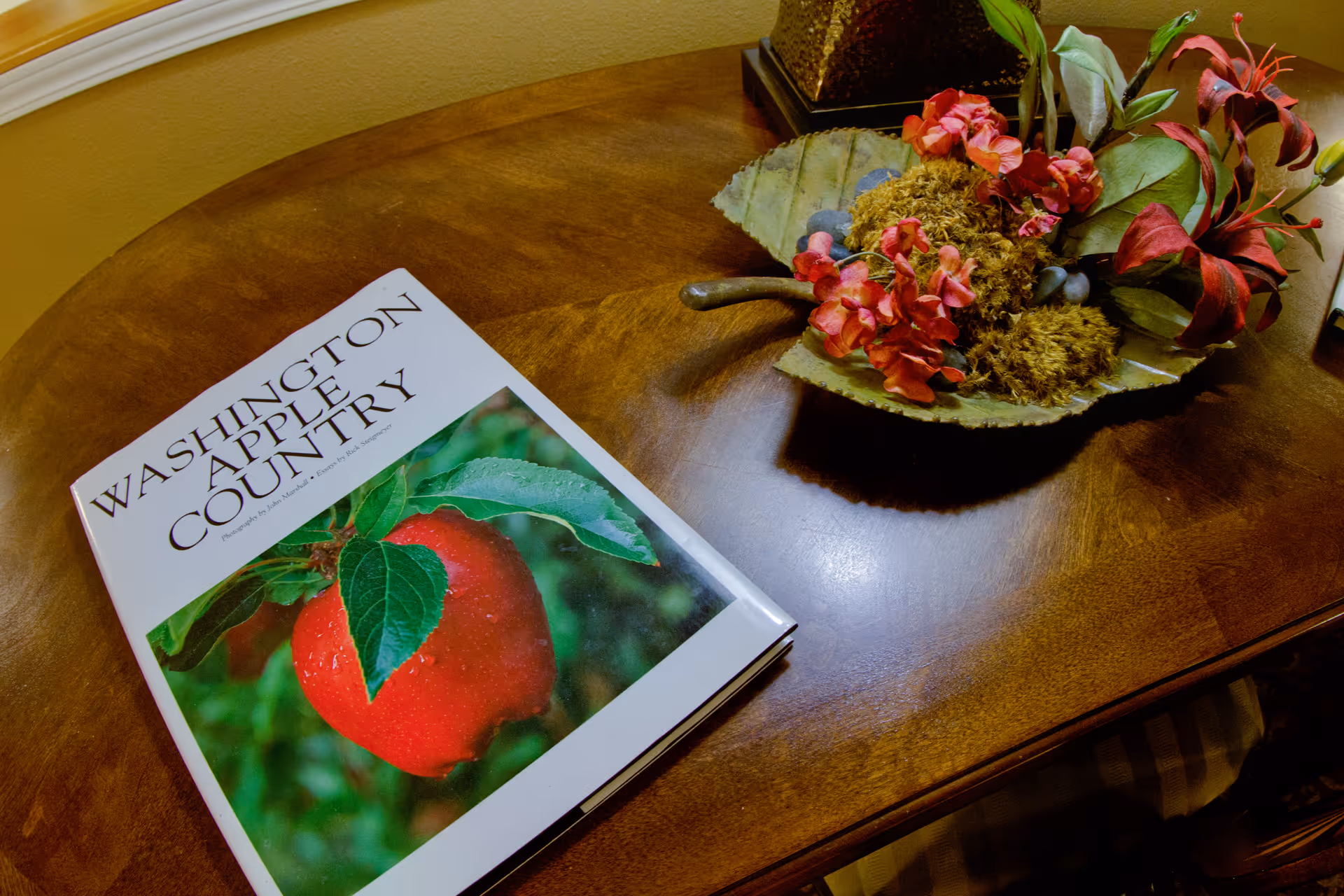 A wooden table with a book titled 'Washington Apple Country' featuring a close-up image of a red apple on the cover, and a decorative floral arrangement with red flowers and green leaves in a leaf-shaped dish.