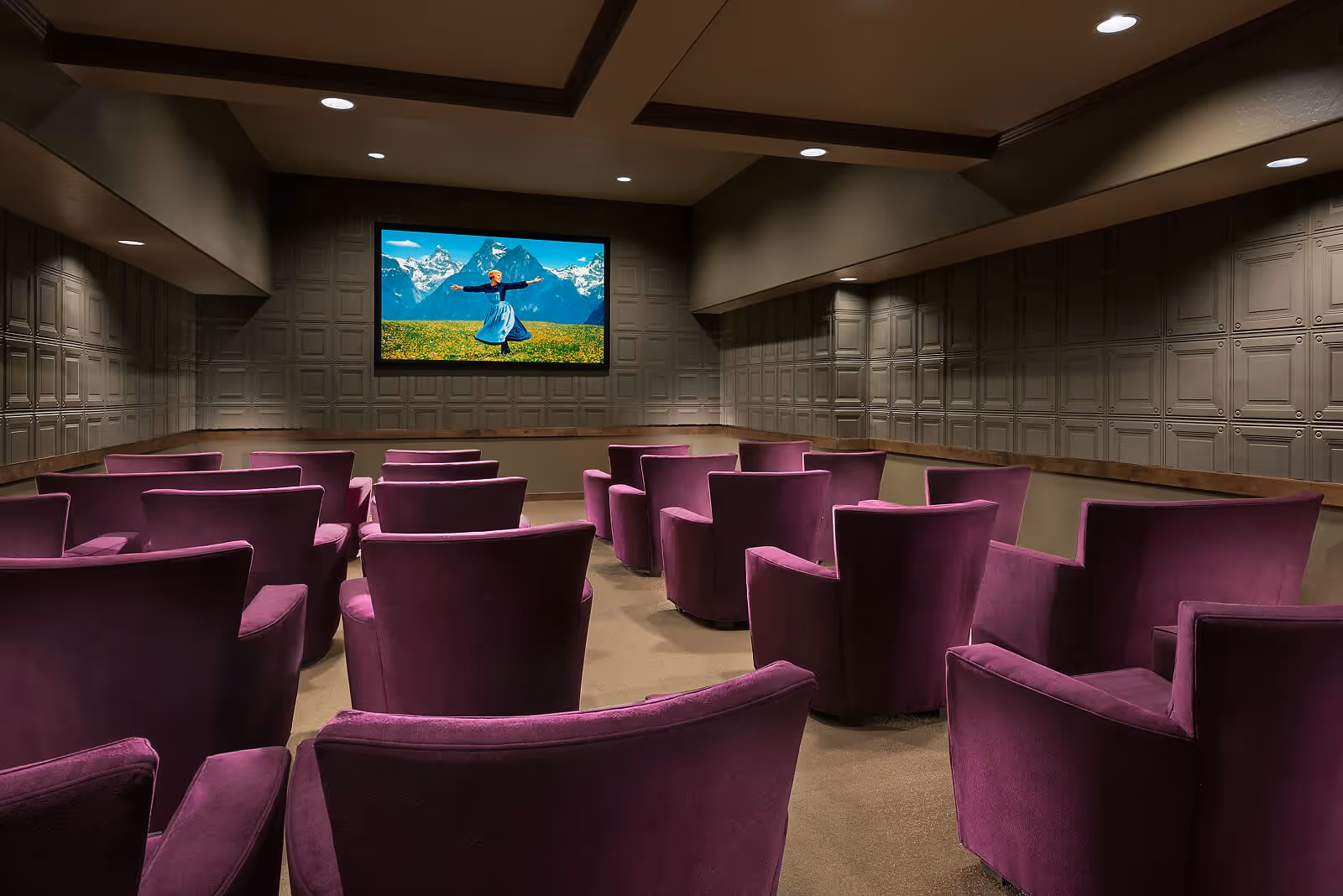 A small theater room with multiple rows of purple upholstered armchairs facing a large screen showing a scene from a movie with mountains and a person dancing in a field.