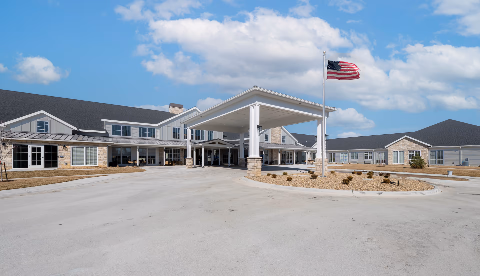 Front exterior view of Cedarhurst Senior Living of Salina building with a covered entrance, an American flag on a flagpole, and a clear blue sky with some clouds.