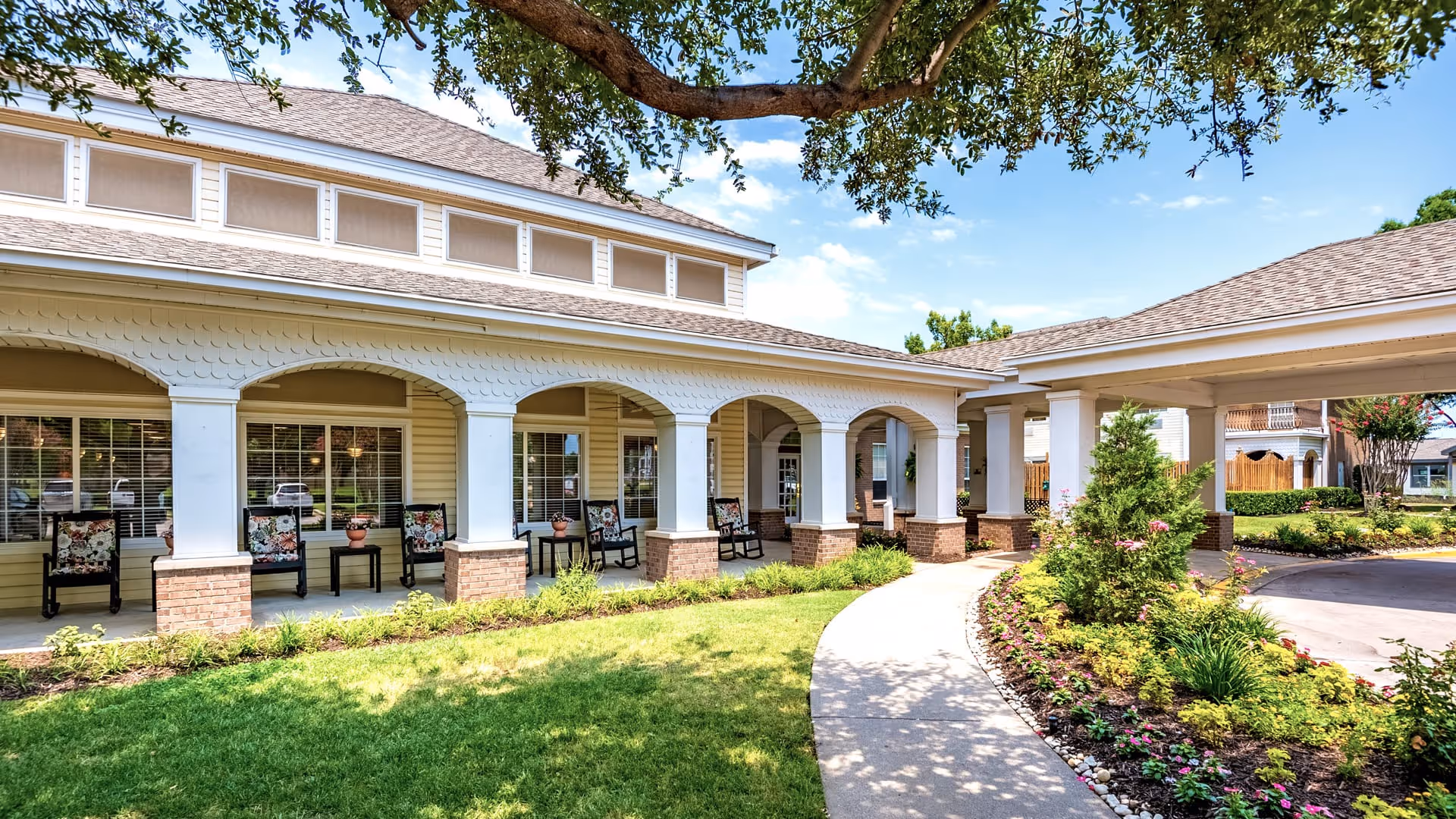 Exterior view of Atria Carrollton senior living facility showing a covered porch with rocking chairs and small tables, surrounded by well-maintained landscaping with green grass, flowering plants, and a curved walkway under a clear blue sky.