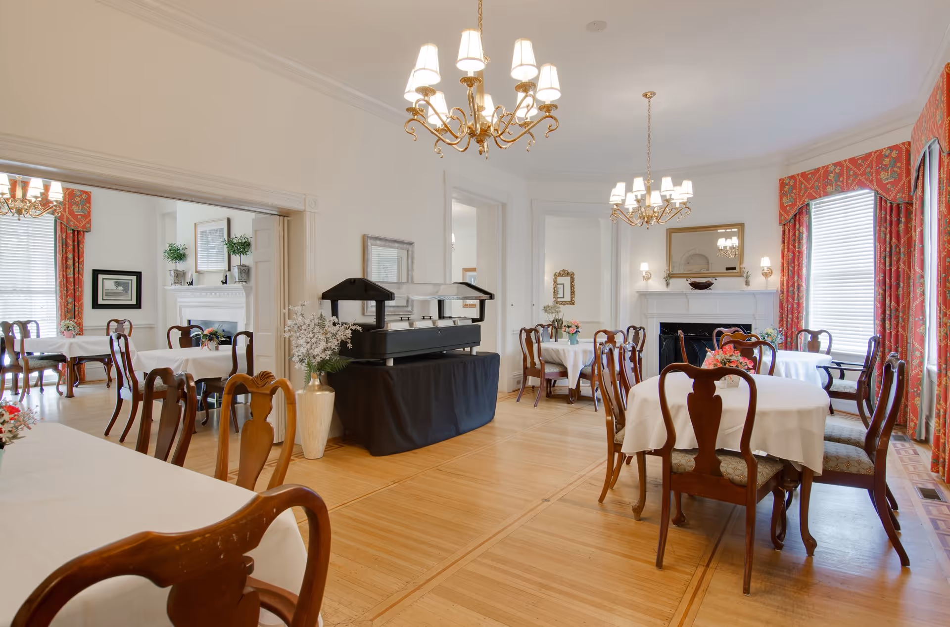 A bright dining room with wooden floors and multiple round tables covered with white tablecloths. Each table is surrounded by wooden chairs with cushioned seats. The room features two ornate chandeliers hanging from the ceiling, large windows with red patterned curtains, and a white fireplace with a mirror above it. There is a buffet station covered with a black cloth in the center of the room, and decorative plants and flowers are placed around the space.