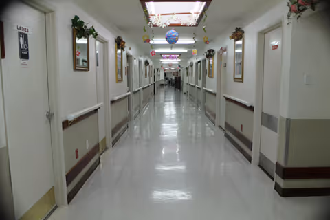 A long, clean hospital hallway with white walls and shiny floors. Doors line both sides of the corridor, some with signs and handrails along the walls. The ceiling has fluorescent lights and decorative hanging ornaments.