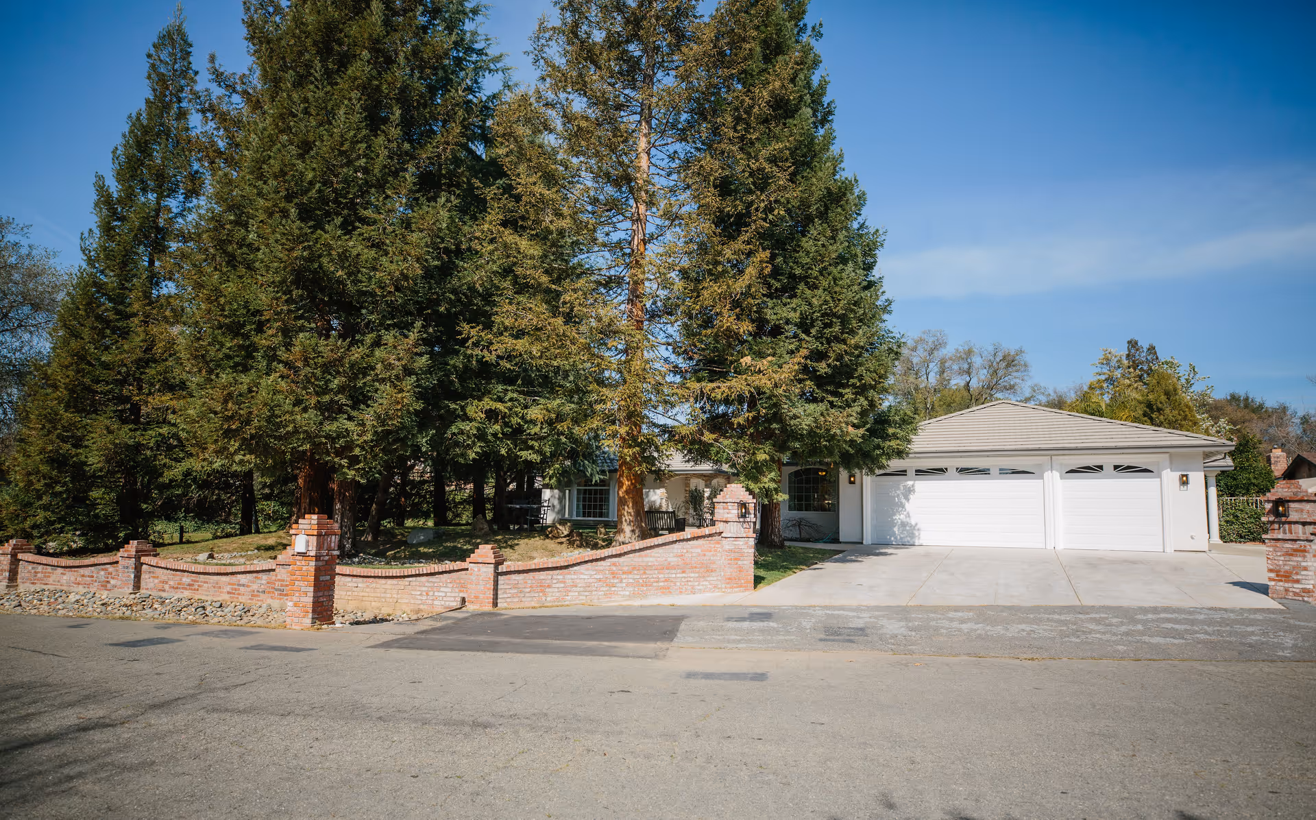 Single-story house with a three-car garage, a low brick front wall, and tall evergreen trees in the front yard.