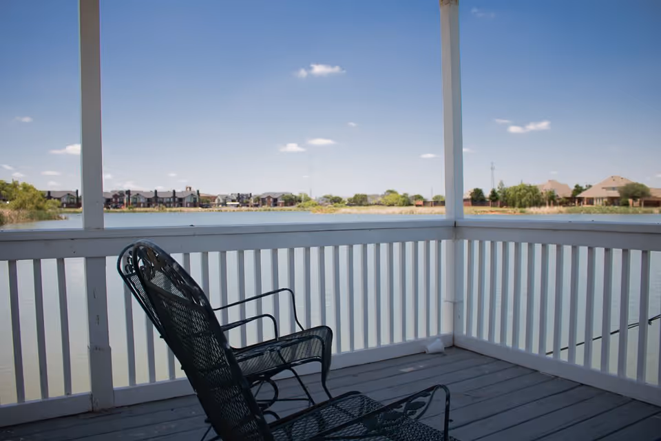 View from a covered porch with white railings overlooking a calm lake, with two black metal chairs facing the water and houses visible in the distance under a blue sky with a few clouds.