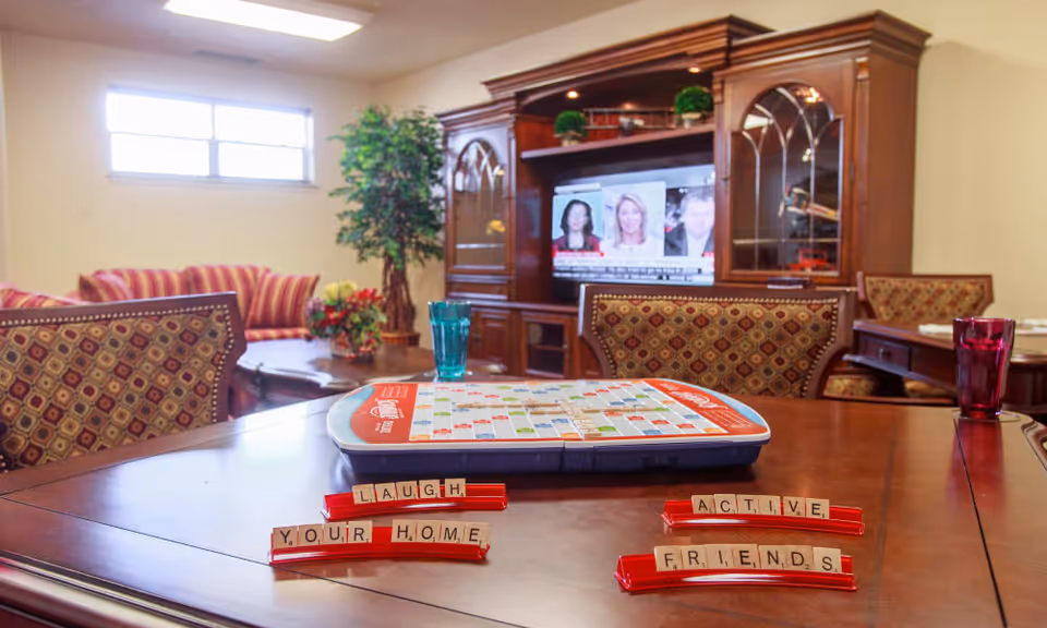A cozy living room area in a senior living facility with a wooden table in the foreground holding a Scrabble game and tiles spelling out 'LAUGH', 'YOUR HOME', 'ACTIVE', and 'FRIENDS'. In the background, there is a wooden entertainment center with a TV showing a news program, a striped sofa, a potted plant, and patterned chairs around the table.