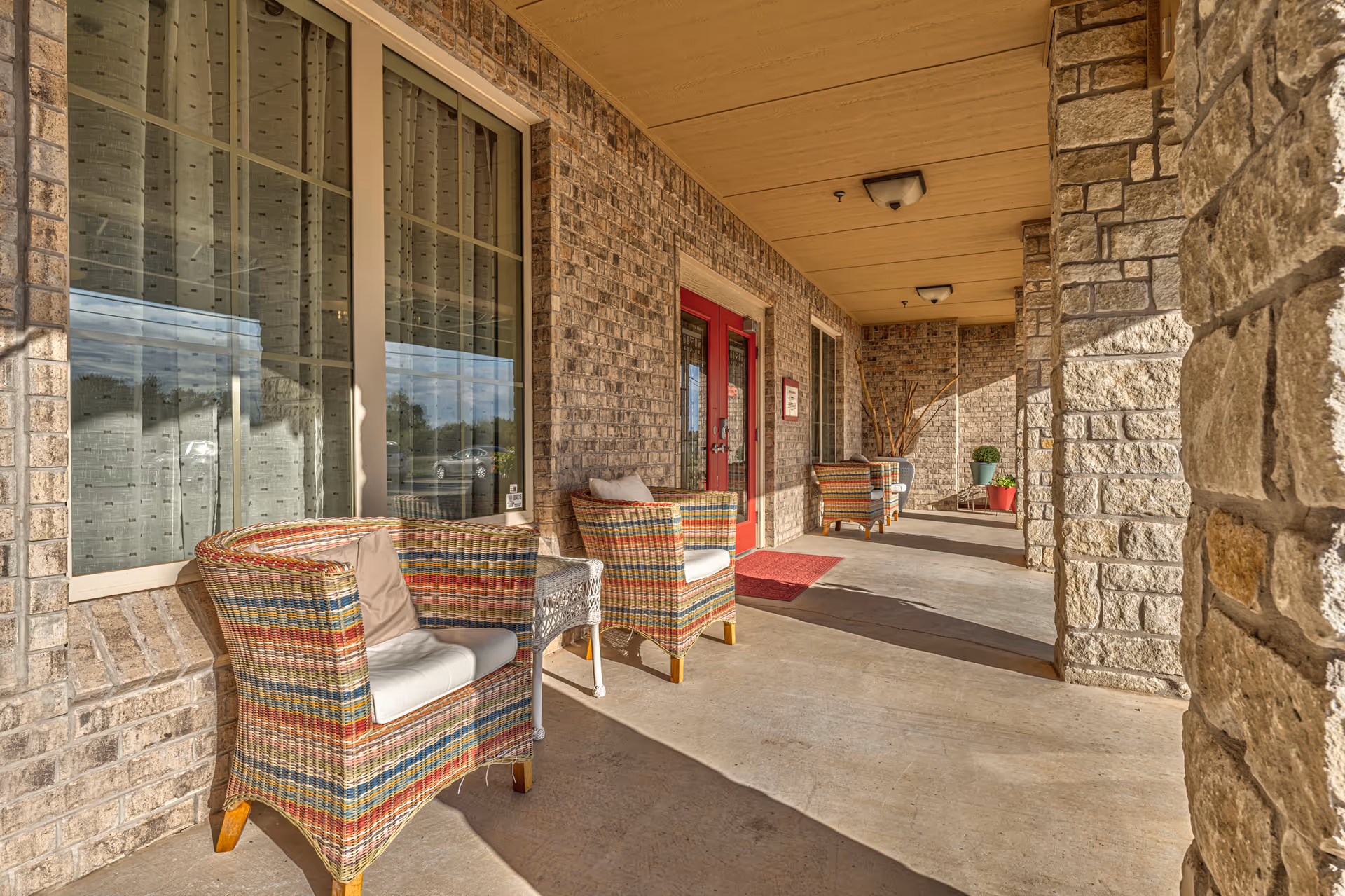 Covered front porch with colorful wicker chairs and a red entrance door at a senior living residence.