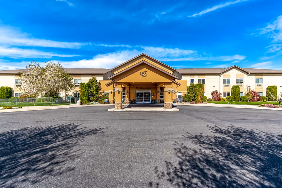 Front exterior view of Ellensburg Senior Living facility with a covered entrance, beige and brown building facade, landscaped bushes and trees, and a clear blue sky.