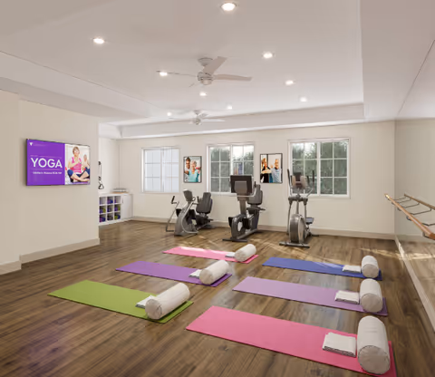 A bright exercise room with colorful yoga mats and bolsters on a wood floor and cardio machines by the windows.