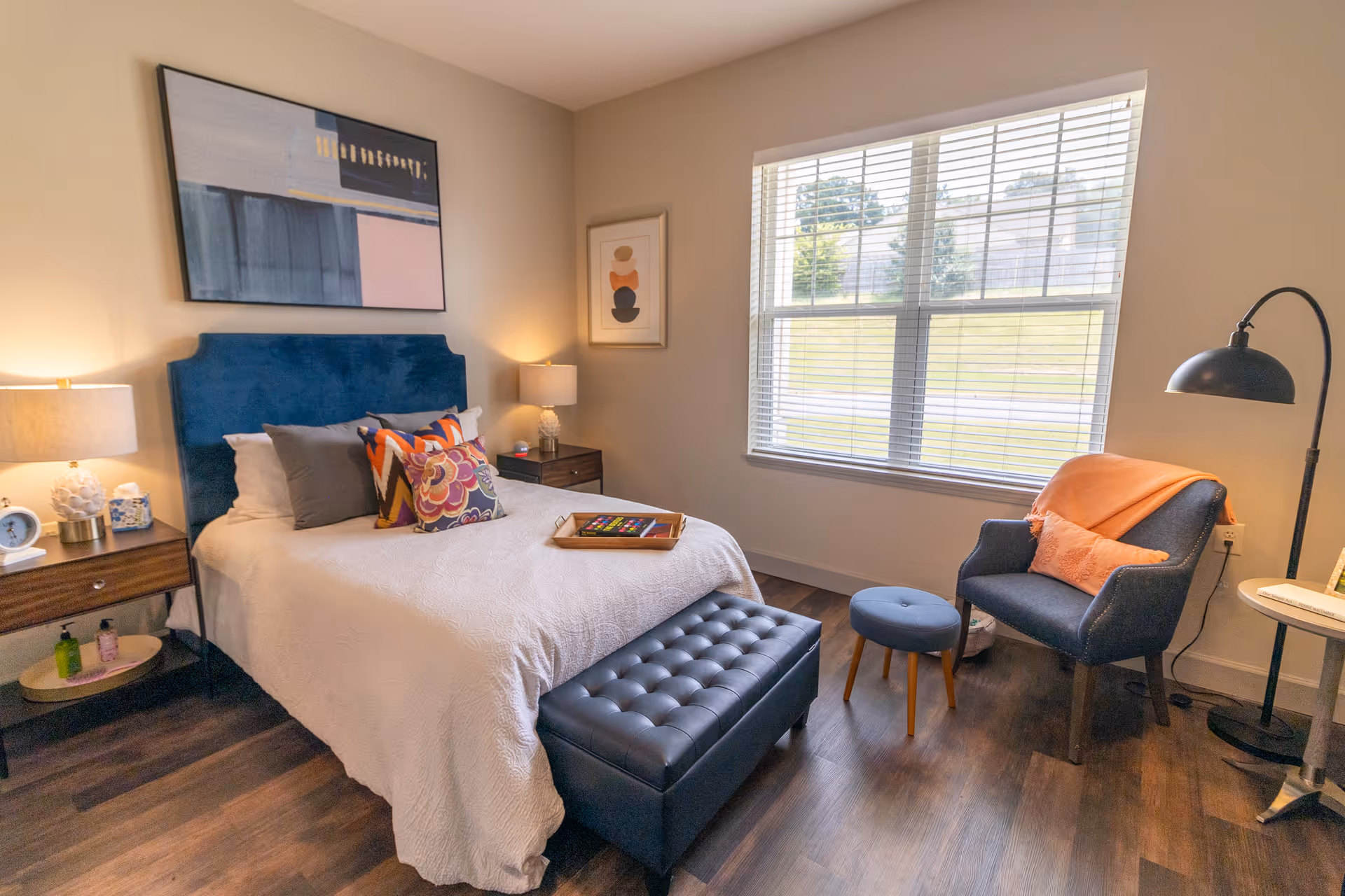 A cozy bedroom with a bed featuring a blue upholstered headboard, white bedding, and colorful decorative pillows. There are two wooden nightstands with lamps on either side of the bed. A large window with blinds lets in natural light. In front of the bed is a black tufted bench. To the right, there is a blue armchair with an orange throw blanket and pillow, a small round side table, and a black floor lamp. The room has wooden flooring and neutral-colored walls with framed artwork.