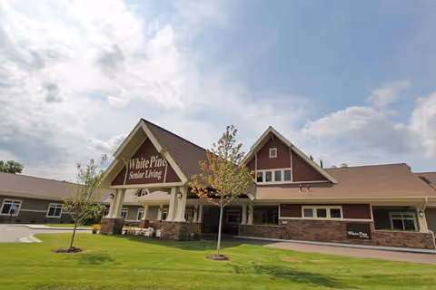 Exterior front view of White Pine Senior Living building with a large covered entrance, green lawn, and a partly cloudy sky.