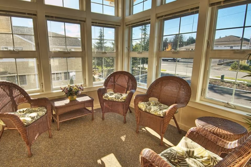 A sunlit sitting area with four wicker chairs featuring patterned cushions arranged around a small wicker table with a potted plant. The space is enclosed by large windows offering a view of neighboring buildings and trees outside.