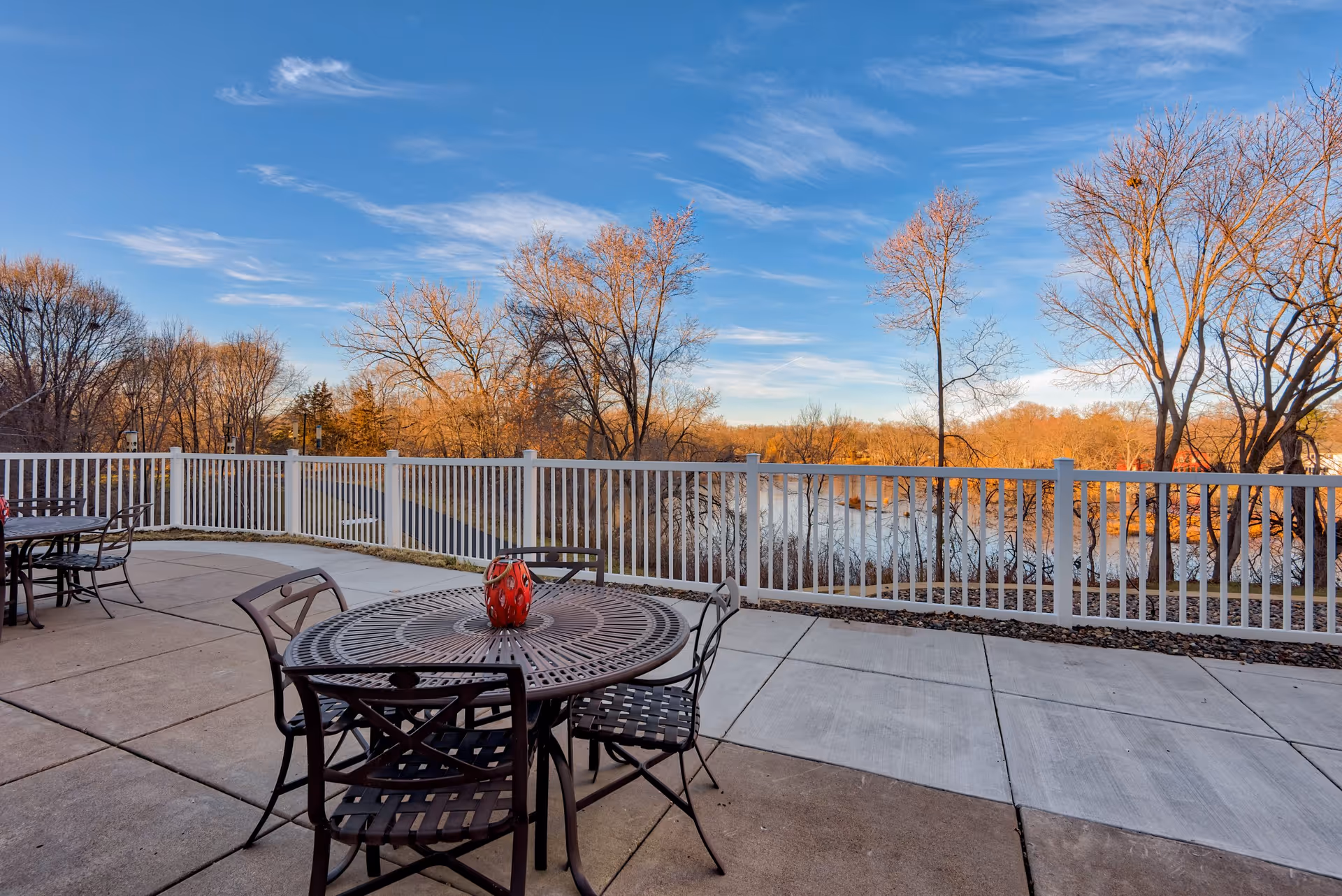Outdoor patio area with metal tables and chairs, a red decorative item on the table, white railing, and trees with autumn foliage under a blue sky.