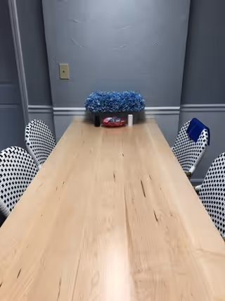 A long wooden table with a light natural finish is centered in a small room with gray walls and white trim. Four white chairs with black geometric patterns are positioned around the table. On the table, there is a blue floral arrangement, a small red dish with items inside, and a white cylindrical object. A light switch is visible on the wall behind the table.