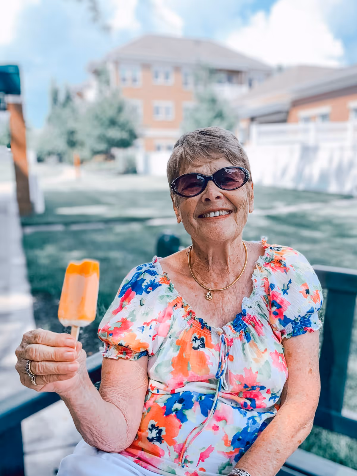 An elderly woman wearing sunglasses and a colorful floral blouse is sitting outdoors on a bench, smiling and holding an orange popsicle. Behind her are green lawns, trees, and brick buildings under a partly cloudy sky.