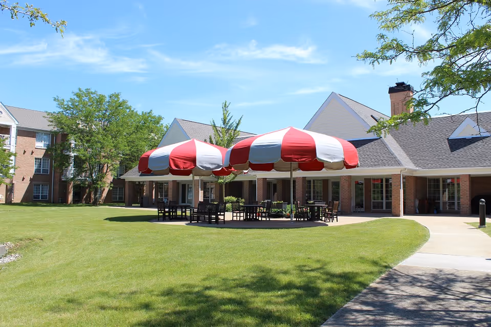 Outdoor patio area at The Parkvue Community featuring two large red and white striped umbrellas shading several tables and chairs on a concrete patio, surrounded by green grass and trees, with a brick building in the background under a clear blue sky.