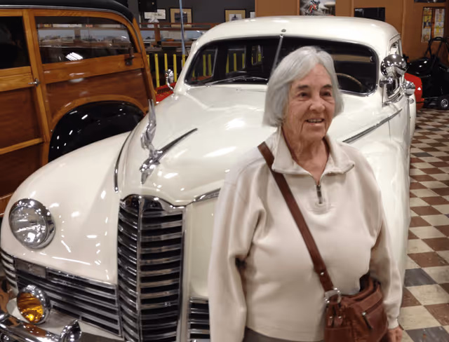 An elderly woman with gray hair wearing a beige sweater and a brown shoulder bag stands smiling in front of a vintage white car inside a building with checkered flooring and other classic cars visible in the background.