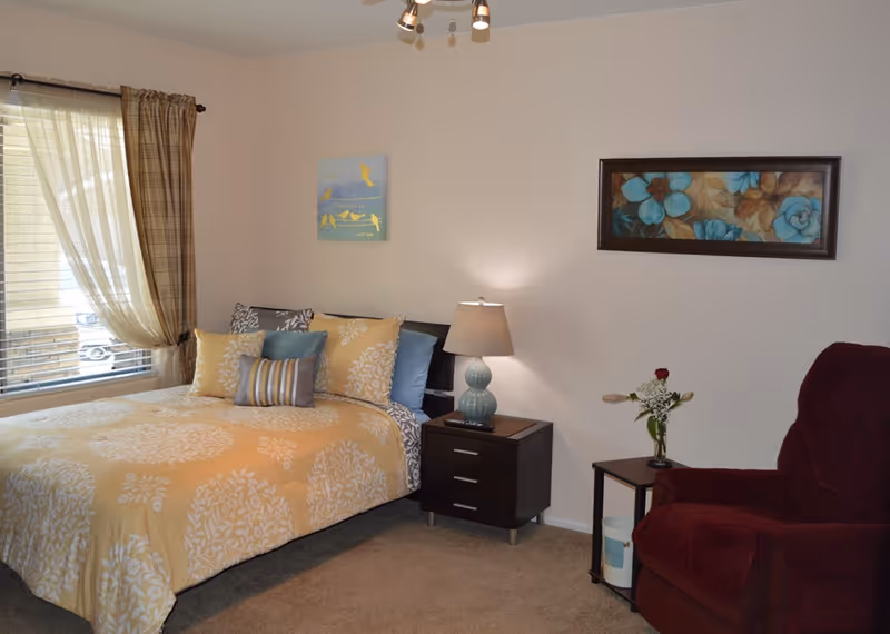 Cozy bedroom featuring a bed with yellow-patterned bedding, nightstand, lamp, and armchair.