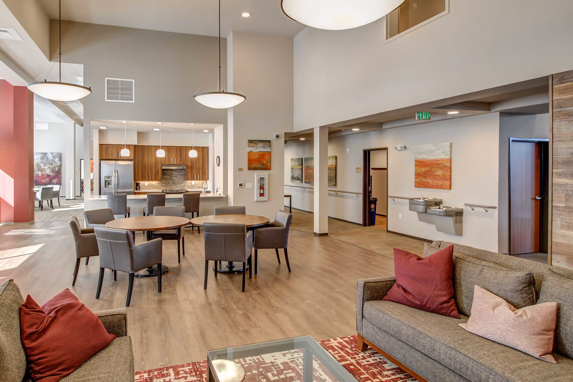 Interior view of a senior living facility common area featuring a seating area with gray couches and red and pink pillows, round tables with chairs, a kitchen area with wooden cabinets and stainless steel appliances, pendant lights, and a hallway with water fountains and artwork on the walls.