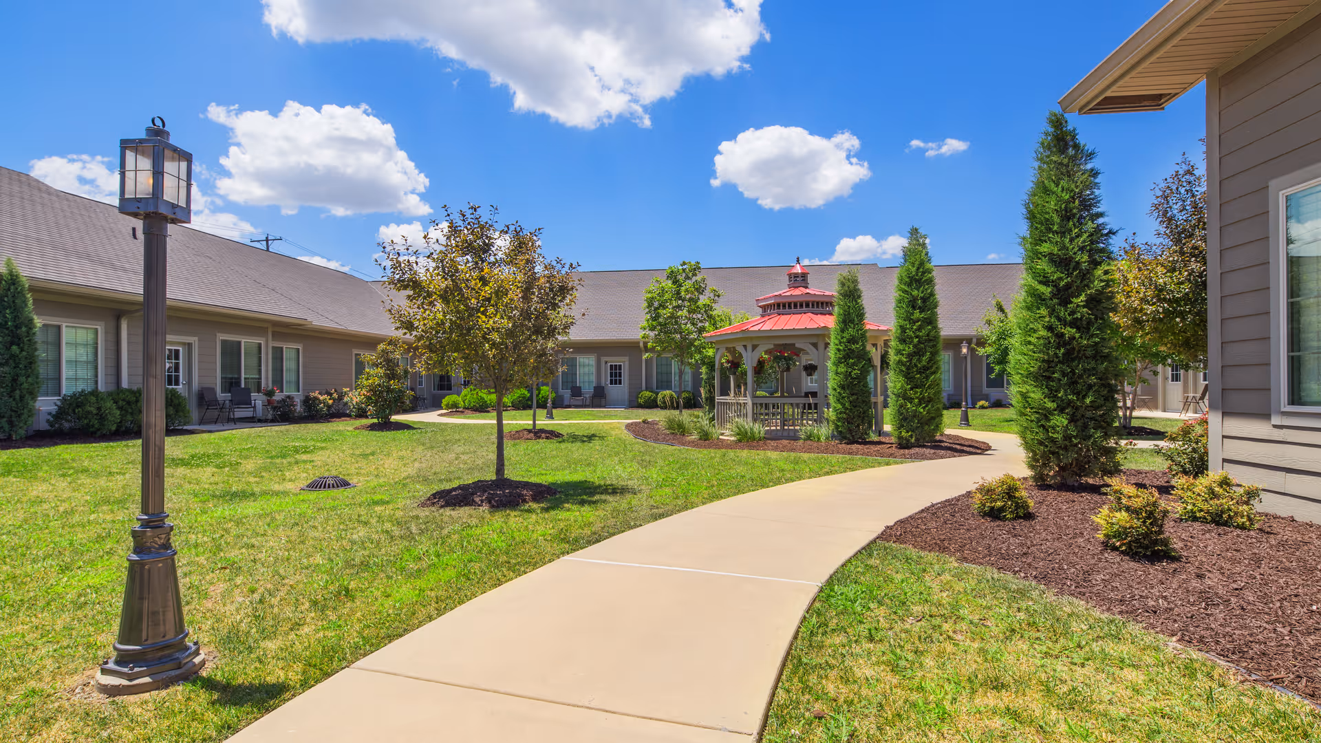 Outdoor courtyard area of Regent Park Assisted Living & Memory Care featuring a curved concrete walkway, green grass, small trees, shrubs, a red-roofed gazebo, and a lamppost under a blue sky with scattered clouds.