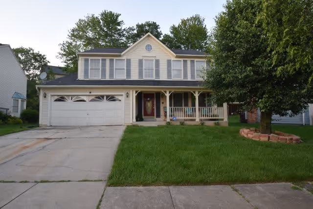 Two-story suburban house with a double garage, a front porch with railings, and a large tree in the front yard surrounded by a circular brick border. The house has beige siding and multiple windows with shutters.