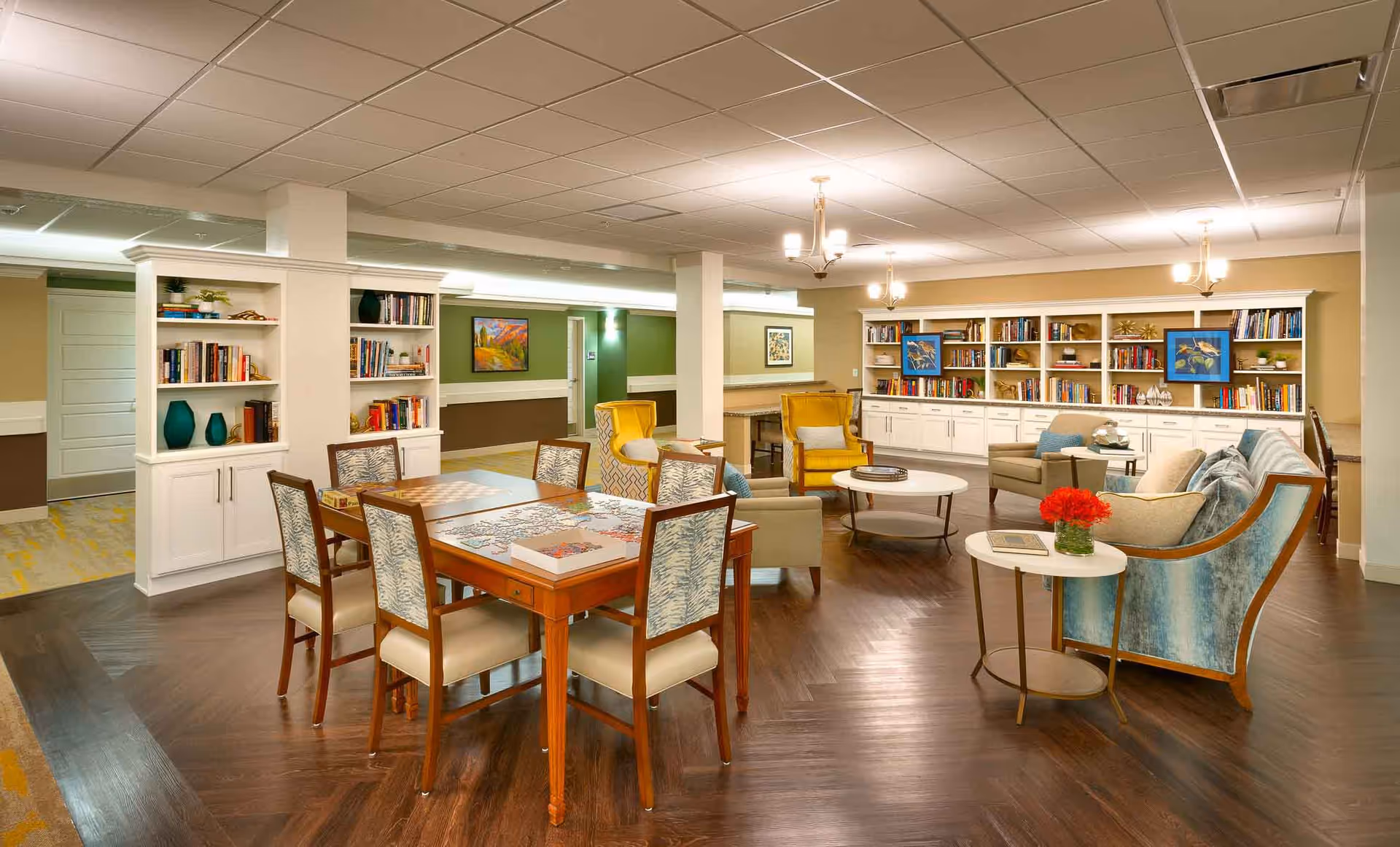 A spacious and well-lit common area in a senior living facility featuring a wooden table with chairs and a puzzle on it, comfortable armchairs and sofas arranged around coffee tables, built-in bookshelves filled with books and decorative items, and warm lighting from ceiling fixtures.