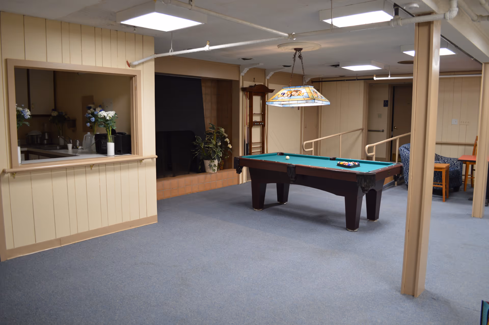 A recreational room with a pool table in the center, a stained glass hanging light above it, a fireplace in the background, and a small seating area with chairs and a table. There is also a window opening to a kitchen area with flowers on the counter.