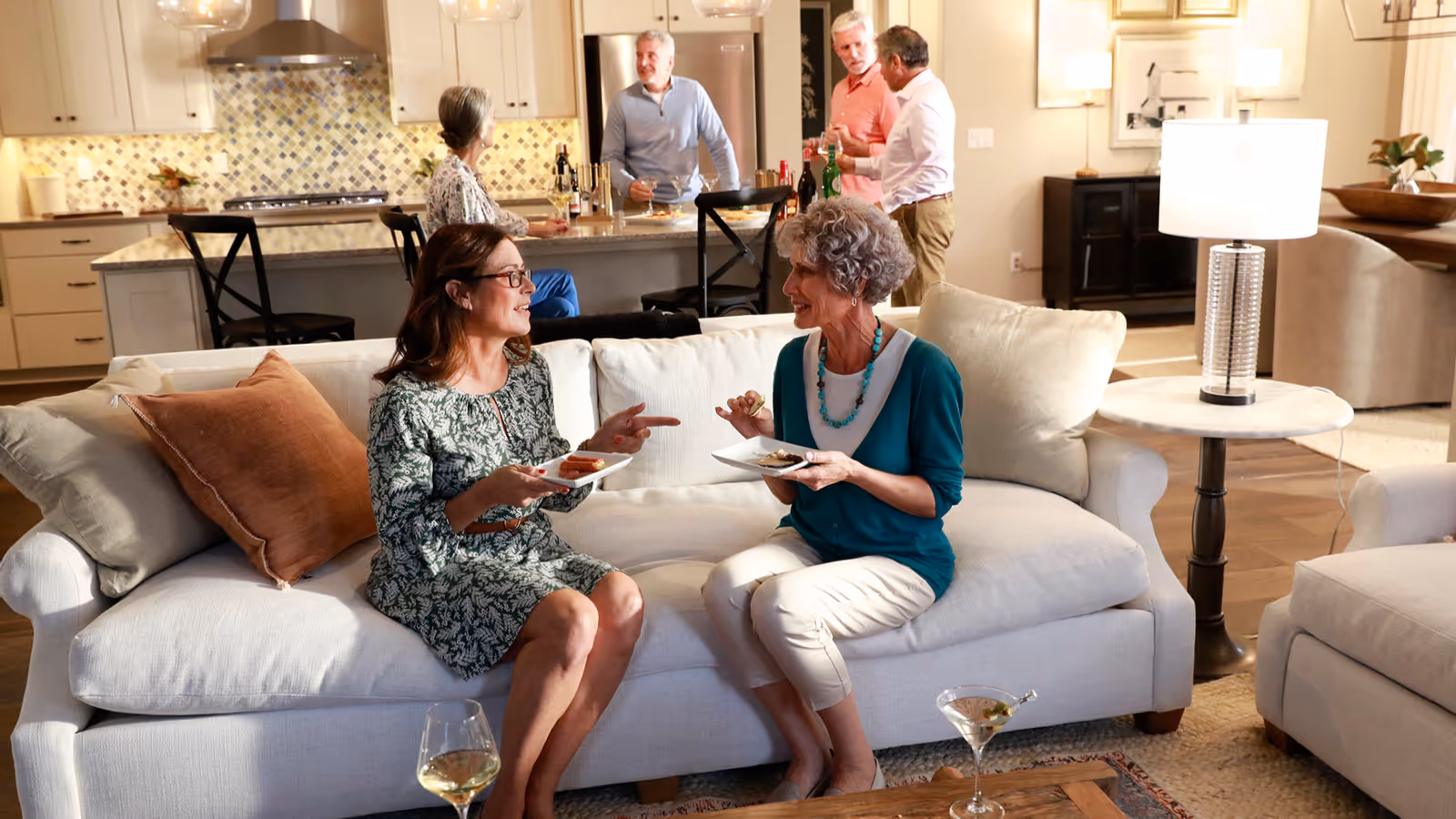 Two women sitting on a white couch in a living room, eating and chatting. In the background, three people stand near a kitchen island with drinks, engaging in conversation. The room is warmly lit with modern decor and a cozy atmosphere.