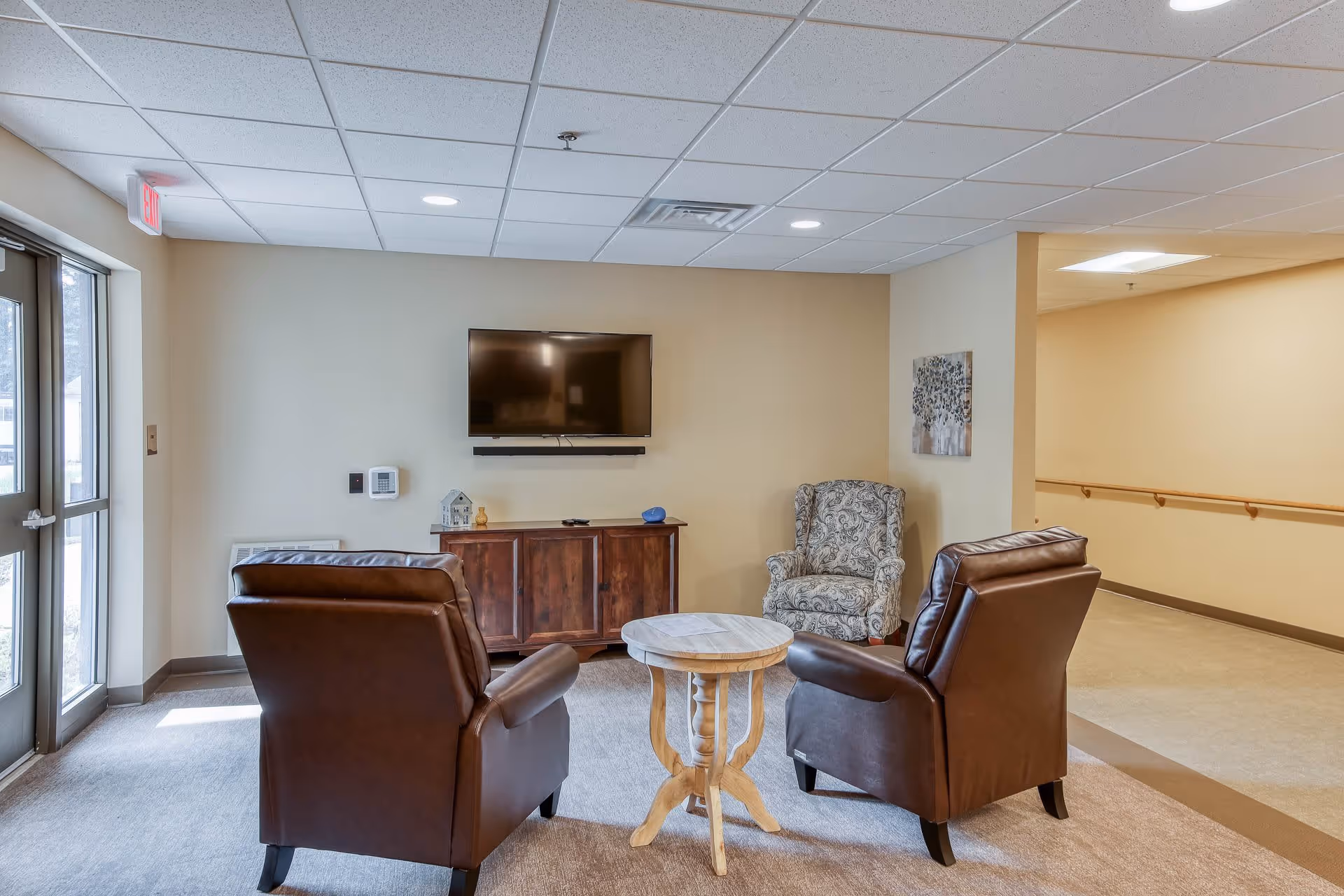 A cozy seating area in a senior living facility with two brown leather armchairs and one patterned armchair arranged around a small round wooden table. A flat-screen TV is mounted on the beige wall above a wooden cabinet. There is a hallway with handrails visible to the right and a glass door to the left letting in natural light.