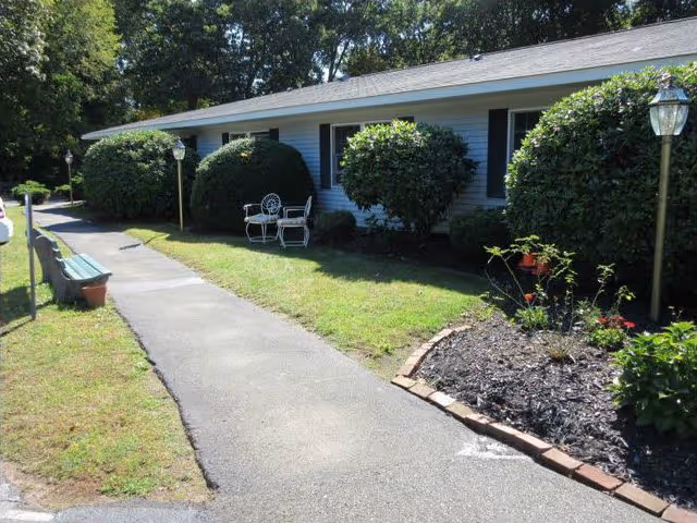 Walkway leading past a single-story retirement home's front with trimmed bushes, outdoor lamps, and a bench.