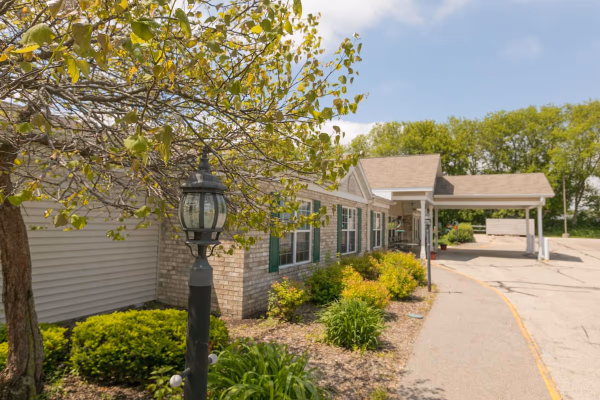 Front entrance of a single-story assisted living building with a covered drop-off, lamp post, and landscaped shrubs.