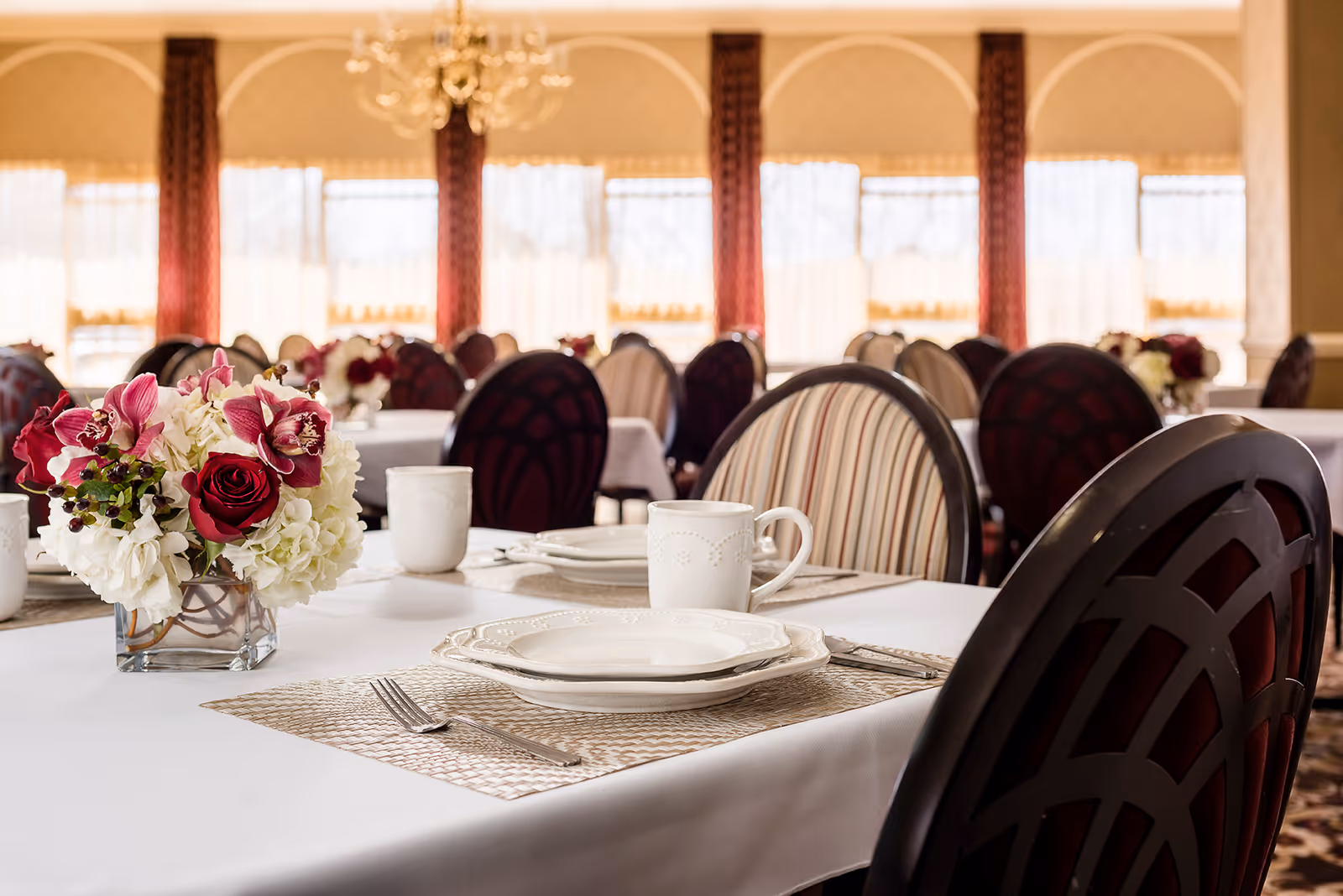 A dining room with tables set for a meal, featuring white tablecloths, white plates, cups, silverware, and floral centerpieces with red and white flowers. The room has large windows with sheer curtains and patterned chairs.