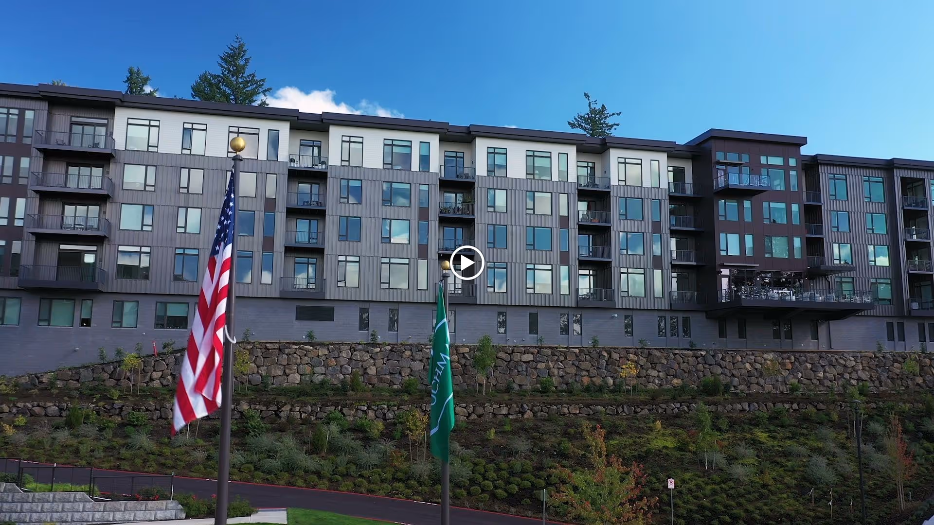 Exterior view of a modern multi-story senior living facility building with balconies, large windows, and a landscaped hillside in front. Two flagpoles with the American flag and a green flag are visible in the foreground under a clear blue sky.